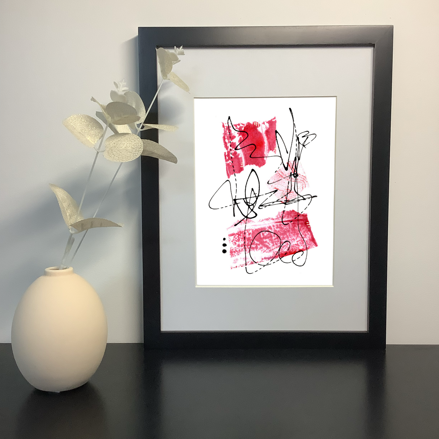 Image featuring an original abstract artwork by artist Julia Hargreaves. The artwork features three pieces of handcrafted collage in shades of red and free flowing line work. It is shown in a black frame on a shelf next to a ceramic vase.