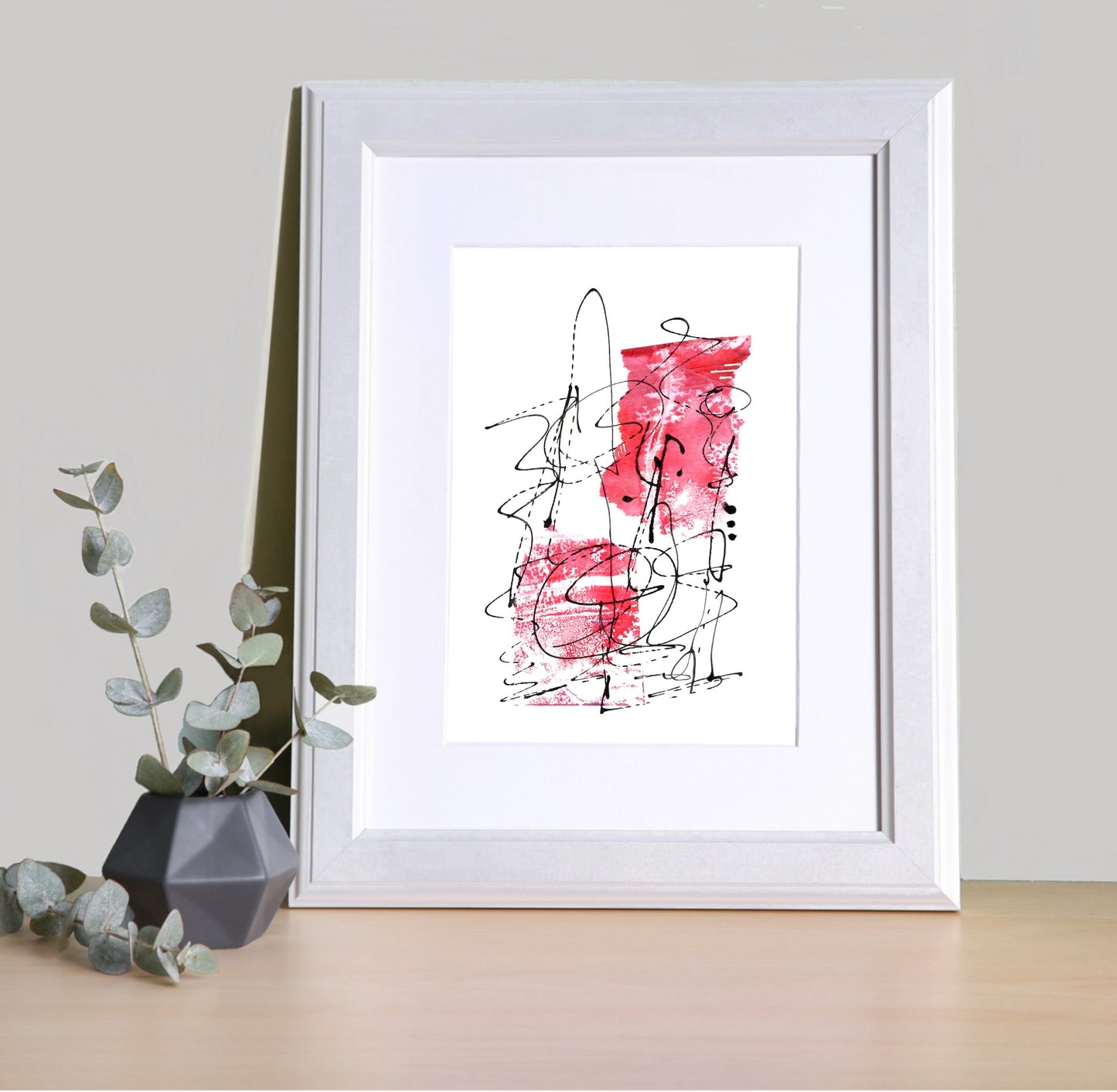 Image featuring a framed abstract artwork on a shelf with a black vase and foliage.  The artwork is abstract in design with red tones and black dynamic line, which give it a lively and expressive vibe.